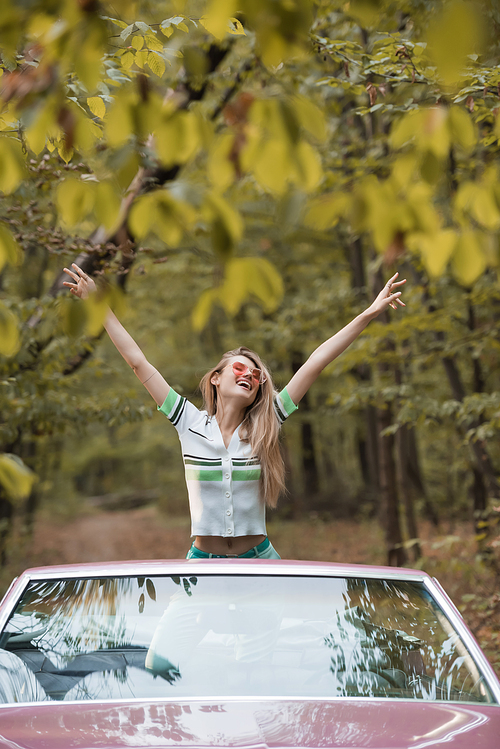 excited woman in sunglasses standing with hands in air in cabriolet