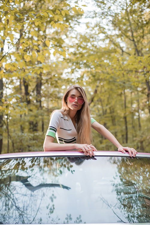 stylish woman in sunglasses  while leaning on windshield of cabriolet