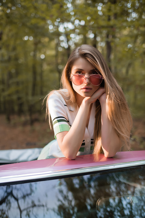 stylish woman in sunglasses holding hands near face while leaning on windshield of cabriolet