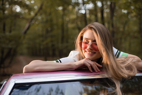 young woman in sunglasses smiling with closed eyes while leaning on windshield of cabriolet