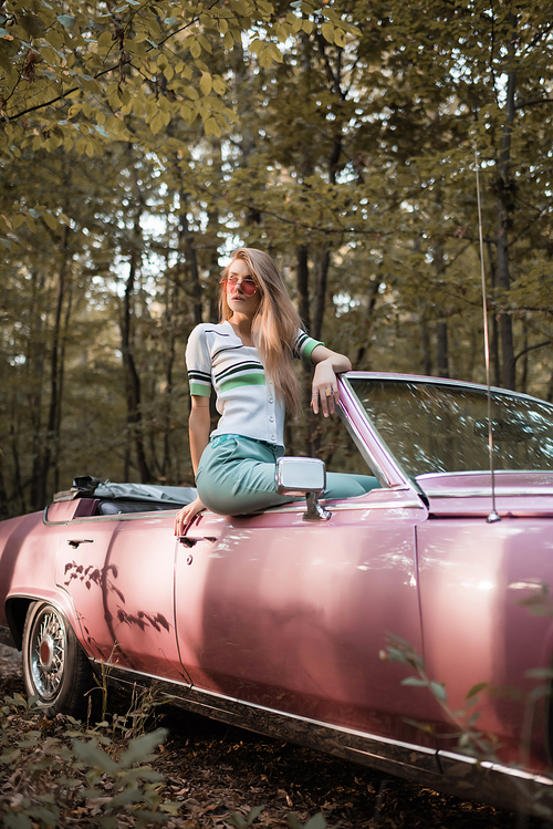 stylish woman in sunglasses looking away while sitting on door of convertible car