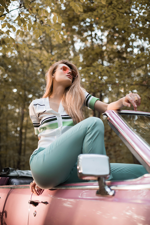 young woman in sunglasses looking away while posing in convertible car on blurred foreground