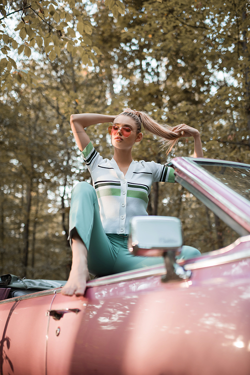 stylish barefoot woman in sunglasses touching hair while posing in cabriolet on blurred foreground