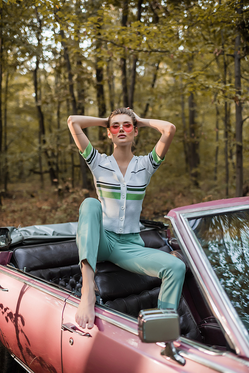 stylish barefoot woman posing in vintage cabriolet with hands behind head