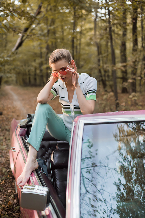 stylish barefoot woman touching eyeglasses while sitting in cabriolet on blurred foreground