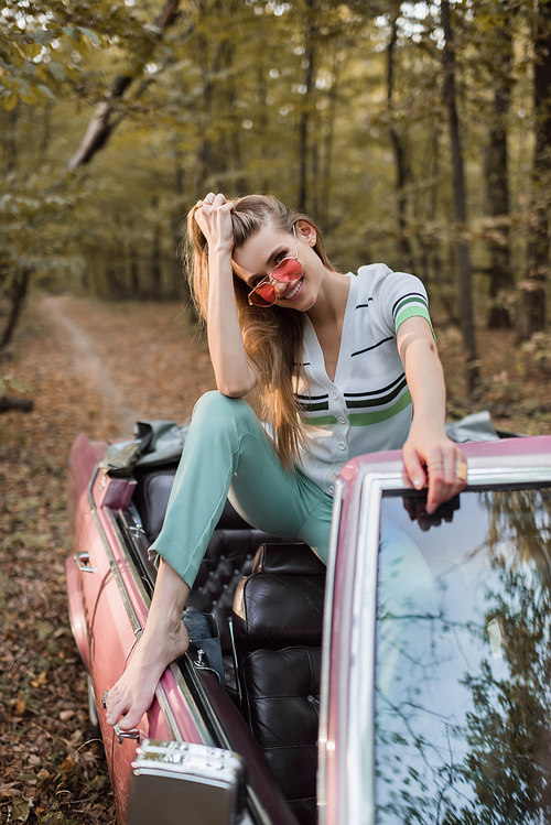 cheerful young woman in sunglasses  while posing in vintage cabriolet on blurred foreground