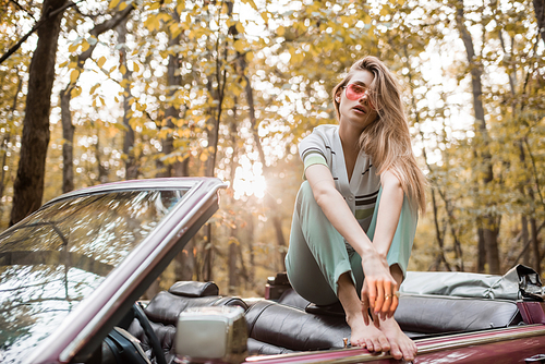 young barefoot woman in sunglasses  while posing in cabriolet on blurred foreground