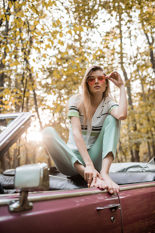 stylish barefoot woman touching sunglasses while posing in cabriolet in forest on blurred foreground