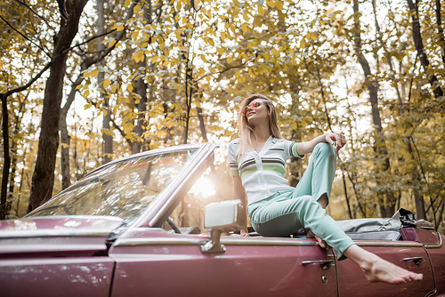 smiling barefoot woman in sunglasses posing in convertible car in forest