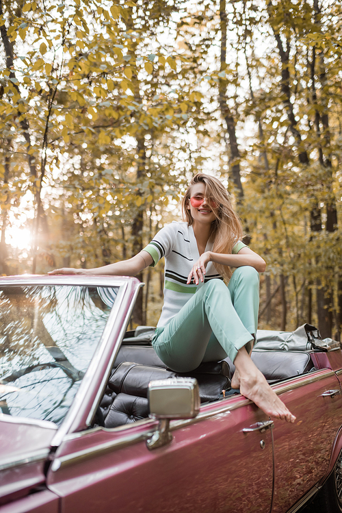 cheerful barefoot woman in eyeglasses  while posing in cabriolet in forest
