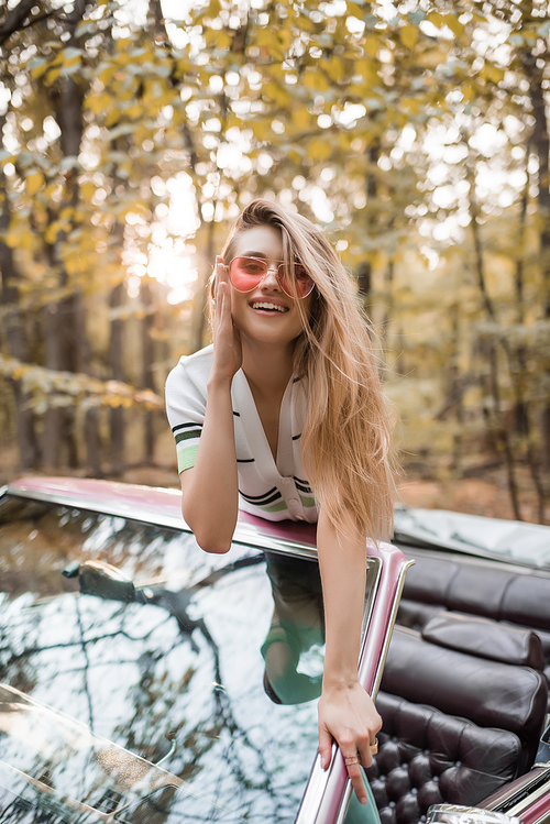 smiling woman touching sunglasses while leaning on windshield of cabriolet and 