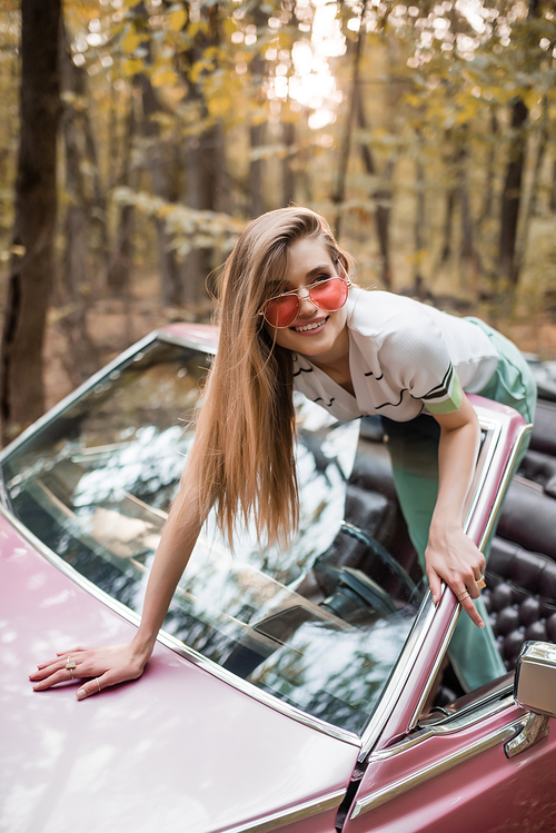 happy woman in sunglasses looking away while leaning on windshield of cabriolet