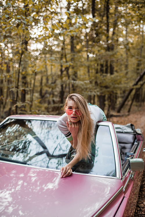 young woman in sunglasses  while leaning on windshield of cabriolet