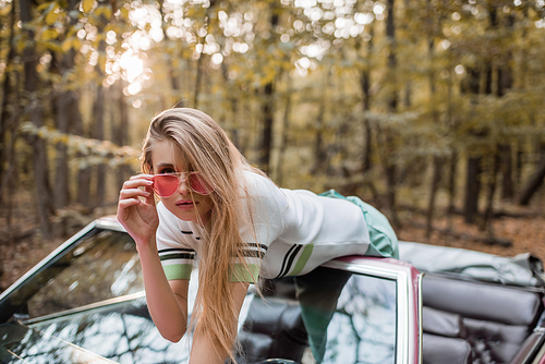 stylish young woman touching sunglasses while leaning on windshield of cabriolet