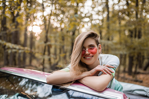 joyful woman in sunglasses  while leaning on windshield of cabriolet with crossed arms