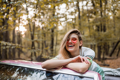 excited woman in sunglasses  while leaning on windshield of cabriolet