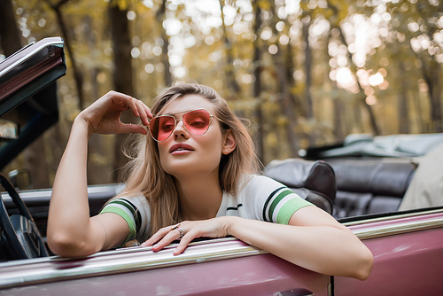 stylish woman touching sunglasses while sitting in cabriolet and looking away