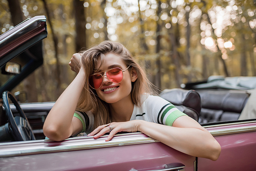 happy woman in sunglasses holding hand near head while sitting in cabriolet with closed eyes