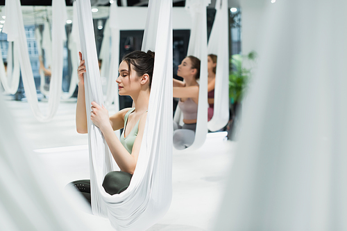 young sportswomen meditating with closed eyes while practicing aerial yoga, blurred foreground