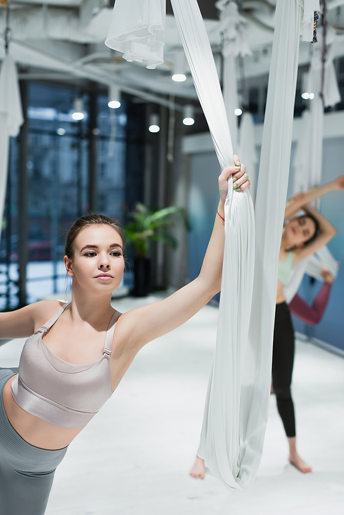 Young woman holding fly yoga straps near sportswomen on blurred background