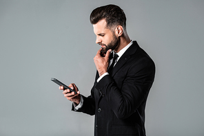 side view of thoughtful businessman in black suit using smartphone isolated on grey