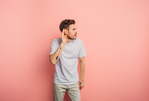 confused young man showing cant hear gesture by holding hand near ear on pink background