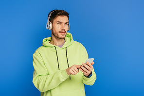 thoughtful young man in wireless headphones using smartphone and looking away isolated on blue