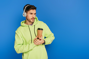 dreamy young man in wireless headphones holding coffee to go and looking away isolated on blue