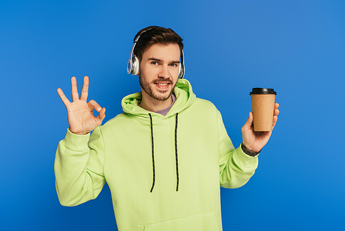 happy young man in wireless headphones holding coffee to go and showing ok sign isolated on blue