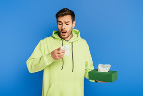 sick man sneezing while holding paper napkins on blue background