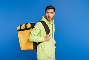 handsome delivery man carrying thermo backpack while looking away isolated on blue