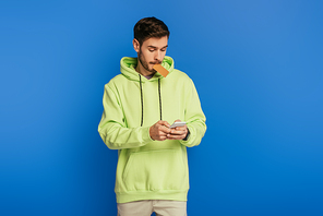 concentrated young man using smartphone while holding credit card isolated on blue