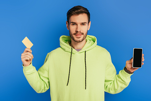positive young man showing smartphone with blank screen and credit card isolated on blue