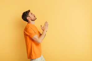 side view of serious young man looking up while praying on yellow background