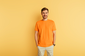 smiling young man in orange t-shirt standing with hands in pockets on yellow background