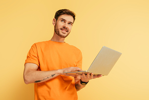 smiling dreamy man looking away while holding laptop on yellow background
