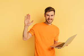 happy man waving hand at camera while holding laptop on yellow background