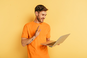 cheerful young man waving hand while having video call on laptop on yellow background
