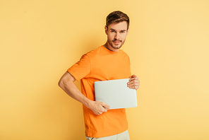 handsome positive man  while holding closed laptop on yellow background