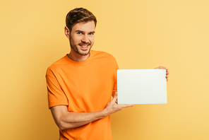 smiling young man showing closed laptop and  on yellow background
