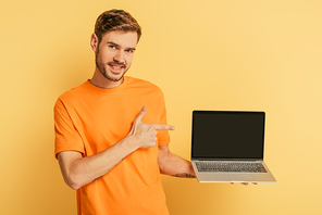 cheerful young man pointing with finger at laptop with blank screen on yellow background