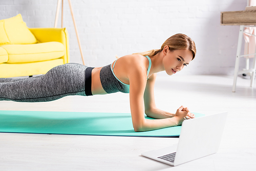 Blonde sportswoman standing in plank near laptop on blurred foreground on floor