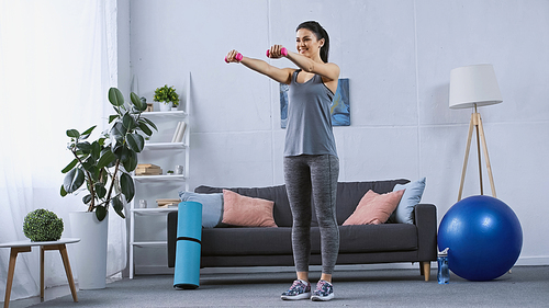 happy young woman in sportswear exercising with dumbbells in modern living room