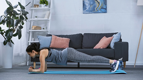 side view of young woman in sportswear doing plank on fitness mat at home
