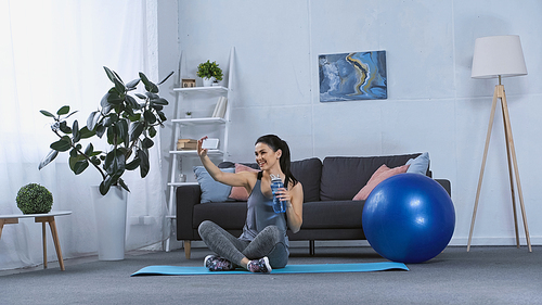 happy young woman in sportswear sitting on fitness mat and taking selfie at home