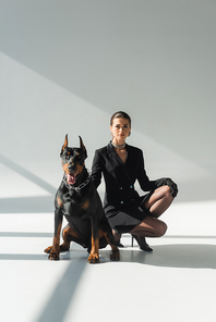 elegant woman in black blazer dress near doberman dog on grey background with shadows