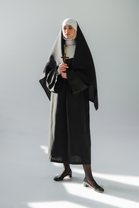 full length view of young nun in black vestment standing with crucifix on grey background