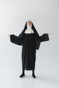 full length view of young nun looking up while praying with open arms on grey background