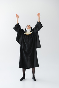 full length view of nun in black vestment praying with raised hands on grey background