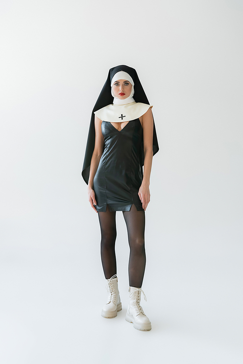 full length view of sexy nun in leather dress and white boots posing on grey